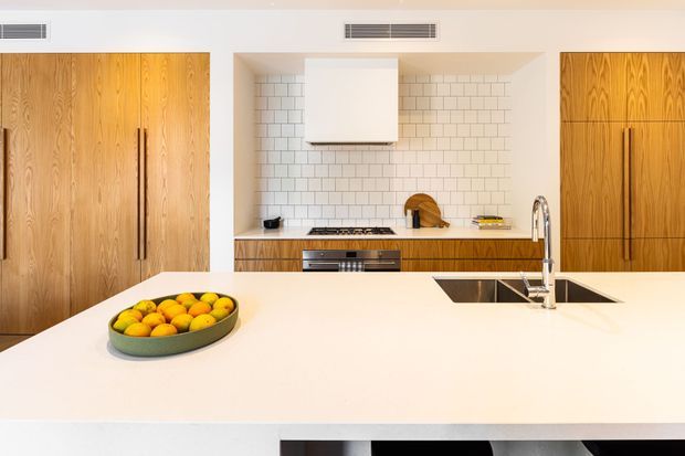 Bright contemporary kitchen featuring extensive warm timber cabinetry, a large white island bench with integrated sink and tapware, a white subway-tile splashback, and concealed appliances. The clean-lined design has a restrained, refined material palette and strong horizontal composition.