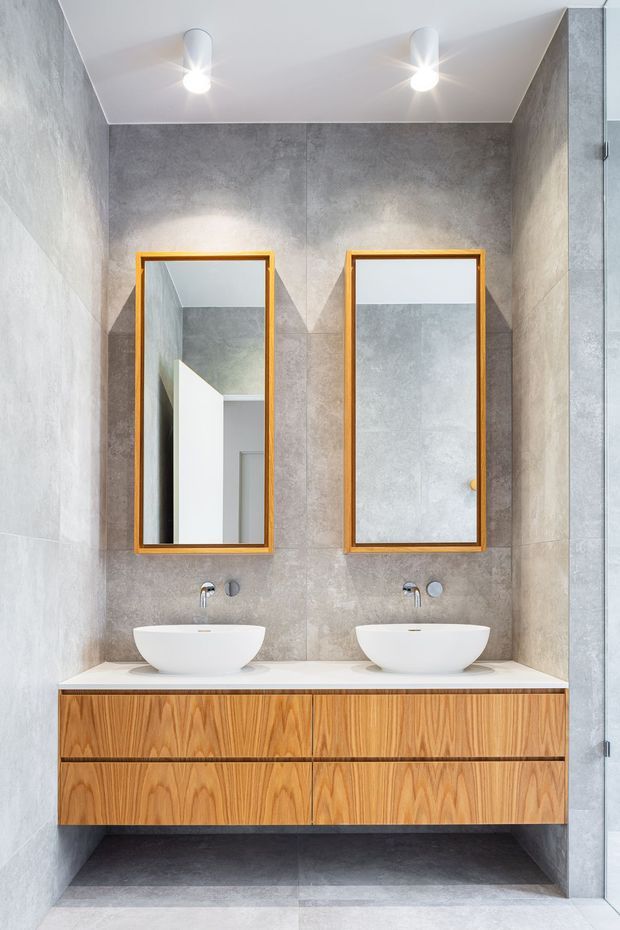 Contemporary double-vanity bathroom with two white vessel basins, wall-mounted chrome taps, a floating timber vanity with integrated drawers, and twin rectangular mirrors with warm wood frames. Large grey concrete-look wall and floor tiles create a neutral, refined finish, complemented by recessed ceiling spotlights and a glass shower enclosure at right.