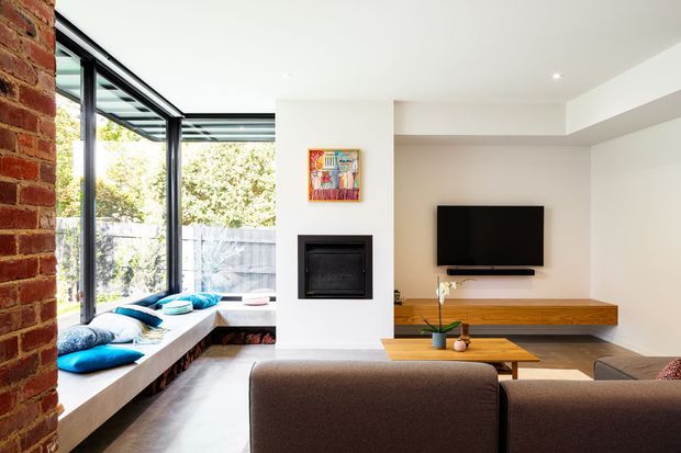 Bright contemporary living room with a wall-mounted television, built-in linear fireplace, and long timber media console. A large corner of black-framed glazing provides views to greenery, while a brick feature wall and built-in window seat with blue cushions add texture and colour. The room includes a brown upholstered sofa and timber coffee table.