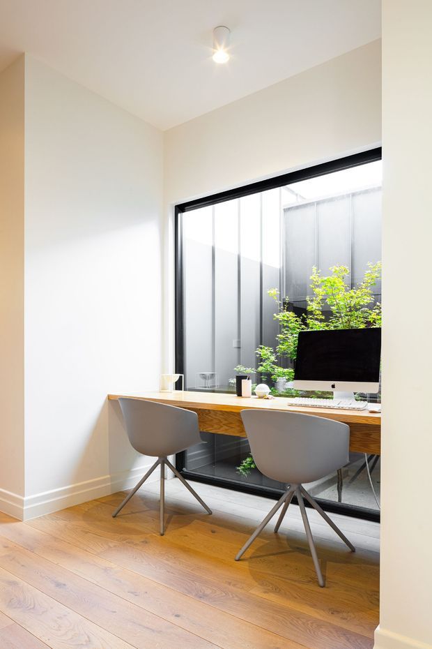 Bright, compact home office with a built-in timber desk positioned in front of full-height black-framed glazing overlooking a planted courtyard. Two modern grey task chairs sit at the desk, which includes a desktop computer and minimal accessories. The room has pale walls, warm timber flooring, and a simple ceiling spotlight.