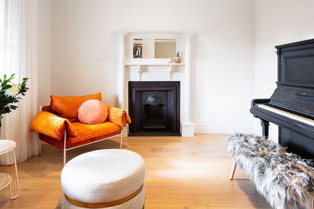 Bright, elegant living room with pale walls, light timber flooring, a restored dark fireplace with white mantel and built-in shelving, an orange upholstered armchair, round ottoman, and black upright piano with a shaggy bench. The room combines classic architectural detailing with colourful contemporary furnishings and abundant natural light.