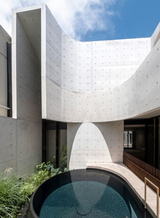 A sculptural interior courtyard enclosed by curved, fair-faced concrete walls with visible form-tie marks. Tall black-framed glazing and a shadowed walkway surround a small curved-edge reflecting pool, with planting along one side and a bright blue sky visible above. The composition is highly minimalist and geometric, emphasizing curved concrete architecture and water.