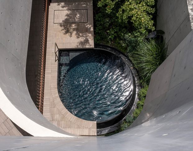 A top-down architectural view of a compact circular swimming pool set within a landscaped courtyard or narrow lightwell. The pool is surrounded by pale timber-look or stone paving, curved dark edging, dense ornamental planting, and tall smooth curved grey walls that create a sculptural enclosed space. Strong sunlight reflects off the rippling blue water.