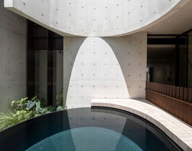 A sculptural contemporary courtyard with a curved dark-tiled pool in the foreground, surrounded by exposed smooth concrete walls and sweeping organic architectural forms. A pale tiled walkway and warm timber-toned vertical balustrade run along the right side, while planted greenery softens the concrete setting.