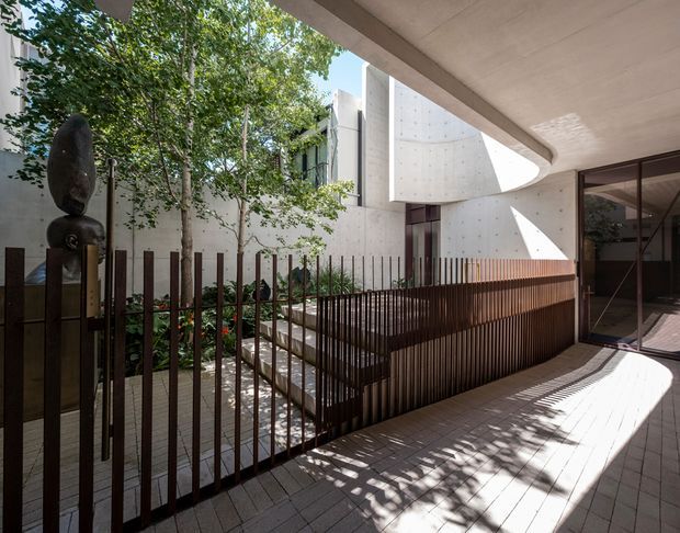 A contemporary covered courtyard or external walkway with a paved floor, dark vertical metal balustrades, timber-toned screening, lush planted garden beds, and sculptural exposed concrete walls. A curved concrete canopy and large oval-like roof opening create dramatic sunlight and shadow patterns. Glazed doors connect to an interior space on the right.