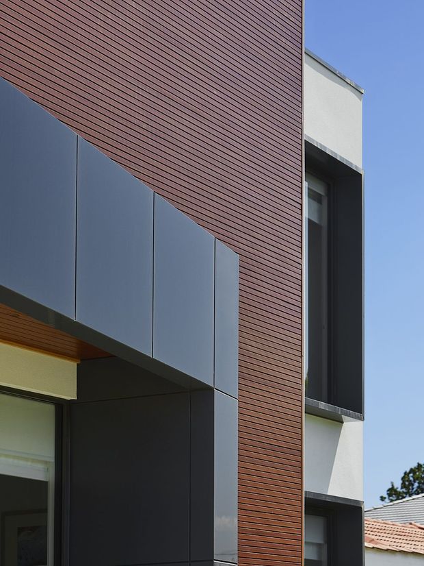 Close-up exterior view of a contemporary multi-level building facade. The composition features dark charcoal panel cladding, a prominent vertical section of narrow reddish-brown timber-look slats, deep-set dark-framed windows, and smooth light rendered walls. Strong geometric lines, projecting volumes, and recessed glazing are highlighted against a clear blue sky.