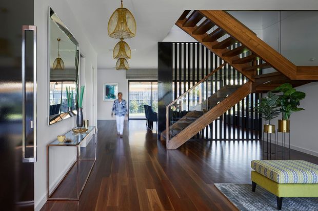 A refined contemporary residential entry and stair hall with dark timber flooring, a sculptural open-riser timber staircase, black vertical balustrade, large wall mirror, slim console, pendant lights with woven shades, and a patterned upholstered bench. Glazing at the rear brings daylight into the circulation space, while a glimpse of adjacent furnishings is visible beyond.