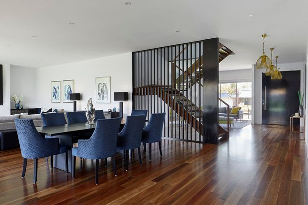 Open-plan contemporary dining and living area with a large dark timber dining table and upholstered navy dining chairs in the foreground. A sculptural timber staircase is screened by a full-height black vertical-slat partition, while polished dark timber flooring extends through the space. White walls, recessed ceiling lights, framed artwork, a dark feature wall and sculptural pendant lights create a refined, high-contrast interior.