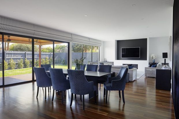 Bright open-plan dining and living area with a large dark timber dining table surrounded by upholstered navy dining chairs. Floor-to-ceiling glazed sliding doors open to a covered outdoor area and garden, while the adjacent living zone includes a pale sofa, media wall with television, and dark cabinetry. The room has polished timber flooring, white walls and ceiling, and a clean contemporary aesthetic.