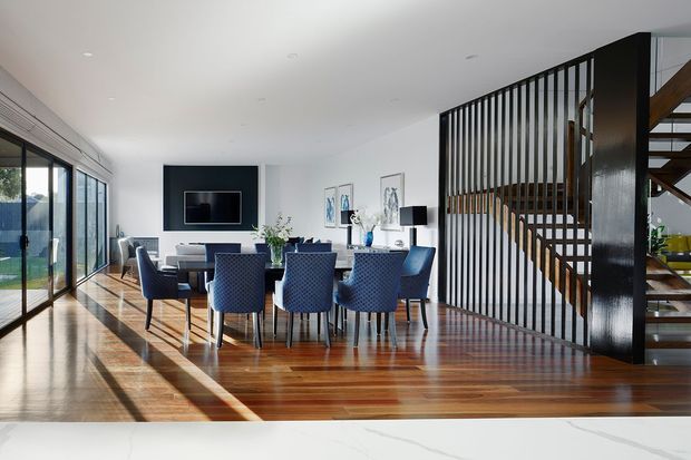 Bright open-plan interior featuring a combined dining and living area. A long timber dining table with blue upholstered chairs sits on polished timber flooring, with a wall-mounted television and contemporary furnishings beyond. Full-height glazed doors line the left side, while a black vertical slatted screen separates the room from an open timber staircase. The space has white walls and ceiling, abundant natural light, and a clean contemporary aesthetic.