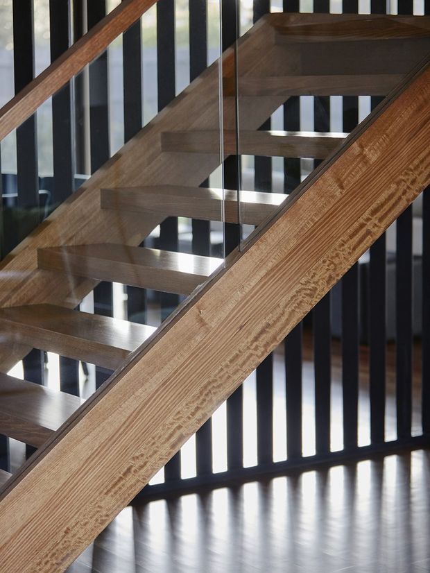 Close-up architectural photograph of a contemporary open-riser staircase with broad timber treads and substantial timber stringers or handrails. Clear glass balustrade panels provide transparency, while a backdrop of evenly spaced black vertical slats creates a strong geometric screen. The polished dark floor reflects light beneath the stair.