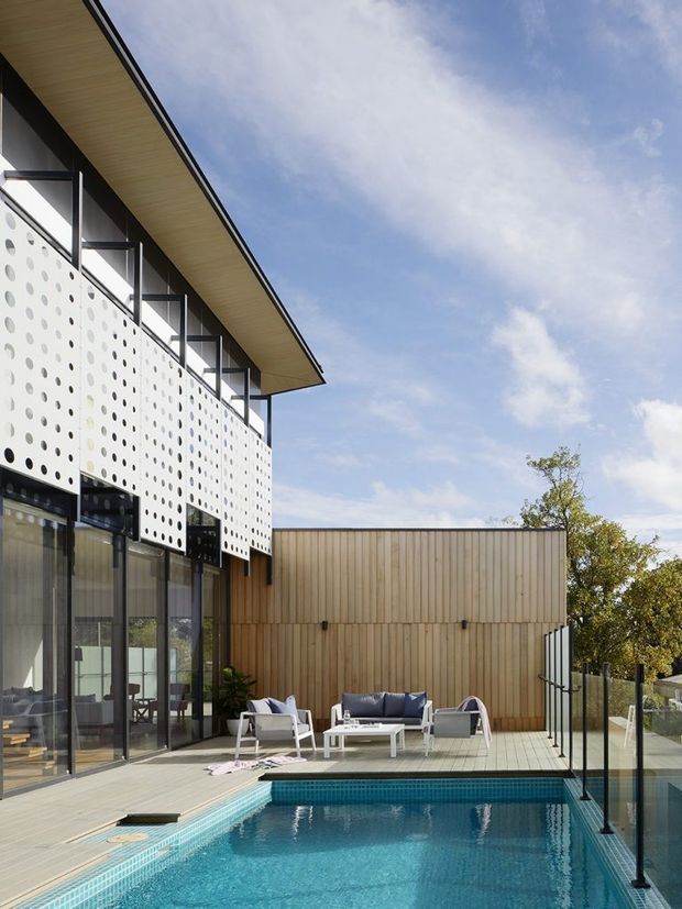 Contemporary multi-level residential exterior overlooking a swimming pool and deck. The building combines a white perforated screen facade, extensive dark-framed glazing, warm vertical timber cladding, and a broad low-pitched roof overhang. Outdoor lounge furniture sits beside the pool, with frameless-style glass pool fencing and a bright open sky.