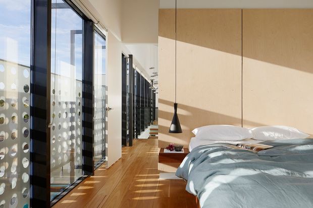 Bright contemporary bedroom with a low platform bed, pale blue bedding, warm timber flooring, and a full-height pale timber feature wall. Black-framed floor-to-ceiling glazing runs along the left side, with a decorative perforated privacy screen visible beyond it. A slim black pendant hangs beside the bed, while strong afternoon sunlight creates geometric shadows across the floor and wall. A corridor or adjoining glazed area extends beyond the bedroom.