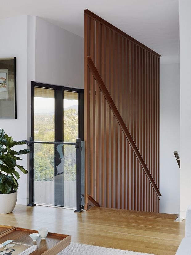 Bright contemporary stairway and landing with a dramatic full-height vertical timber slat screen and integrated diagonal handrail. Pale timber flooring and steps contrast with white walls, while black-framed floor-to-ceiling glazing opens to a leafy outlook. A small seating area and indoor plant are visible in the foreground.