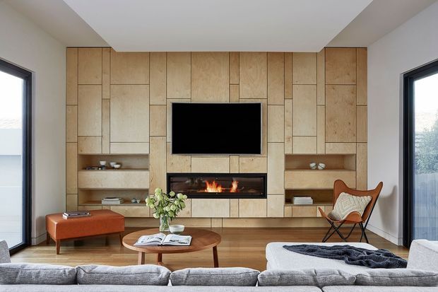 A bright contemporary living room centered on a full-height light timber feature wall with geometric paneling, built-in open shelving, a wall-mounted television, and a linear fireplace. A large pale sectional sofa faces the feature wall, with a warm rust-colored ottoman, round timber coffee table, and leather lounge chair. Floor-to-ceiling glazing on both sides brings in natural light, while timber flooring and restrained furnishings create a refined, welcoming atmosphere.