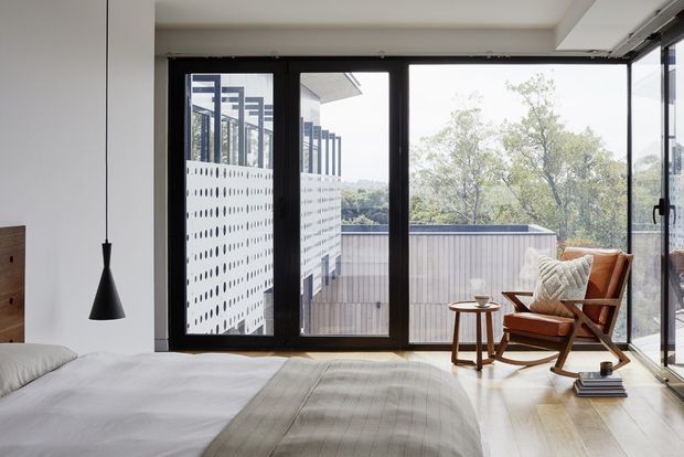 A bright contemporary bedroom with a low-profile bed, pale bedding, a textured neutral runner, and a timber-and-leather lounge chair beside a small round side table. Full-height black-framed glazing opens to a balcony or terrace, with a perforated architectural screen and treetop outlook beyond. A minimalist black pendant hangs beside the bed, and pale timber flooring runs throughout.