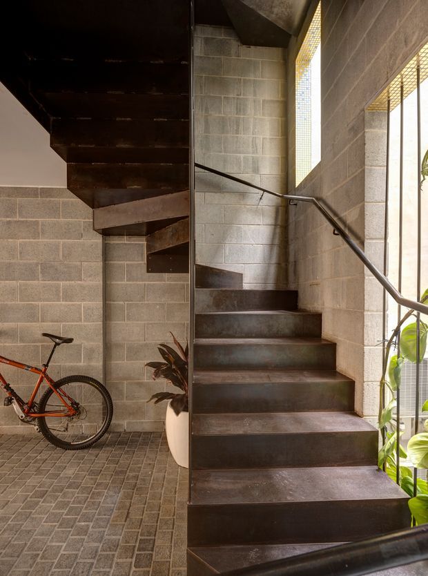 Industrial-style interior stairwell with a dark steel staircase and matching handrail, exposed grey concrete block walls, narrow translucent and glazed openings, and a textured masonry-tile floor. A bicycle and potted plants occupy the lower landing, adding functional and organic elements to the otherwise utilitarian space.