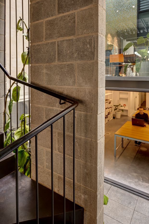 Interior architectural view of a multi-level commercial or office space, focused on a black metal stair and railing beside a tall exposed concrete block wall. Large glazed openings overlook a lower-level workspace with desks, shelving, plants, and people. The space has an industrial, utilitarian character with polished concrete floors and strong vertical circulation.