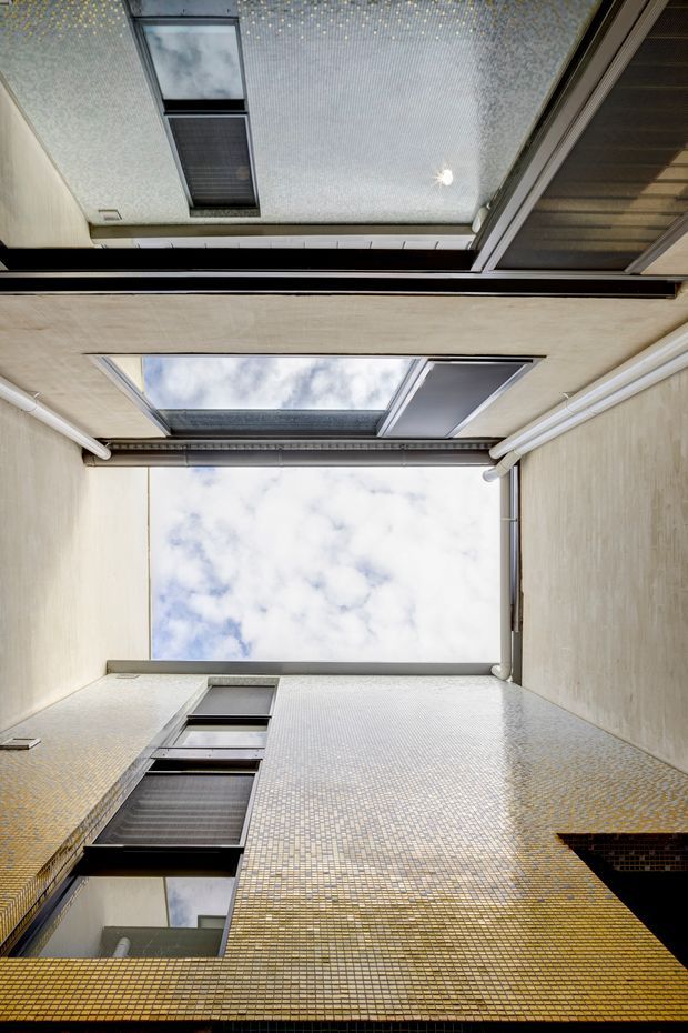 Upward-looking view into a narrow enclosed courtyard or light well, surrounded by tall masonry walls clad in small yellow and cream mosaic tiles. Several dark-framed windows and glazed skylight-like openings overlook the void, with exposed guttering and a bright sky visible above. The image emphasizes the vertical courtyard geometry and reflective glazing.
