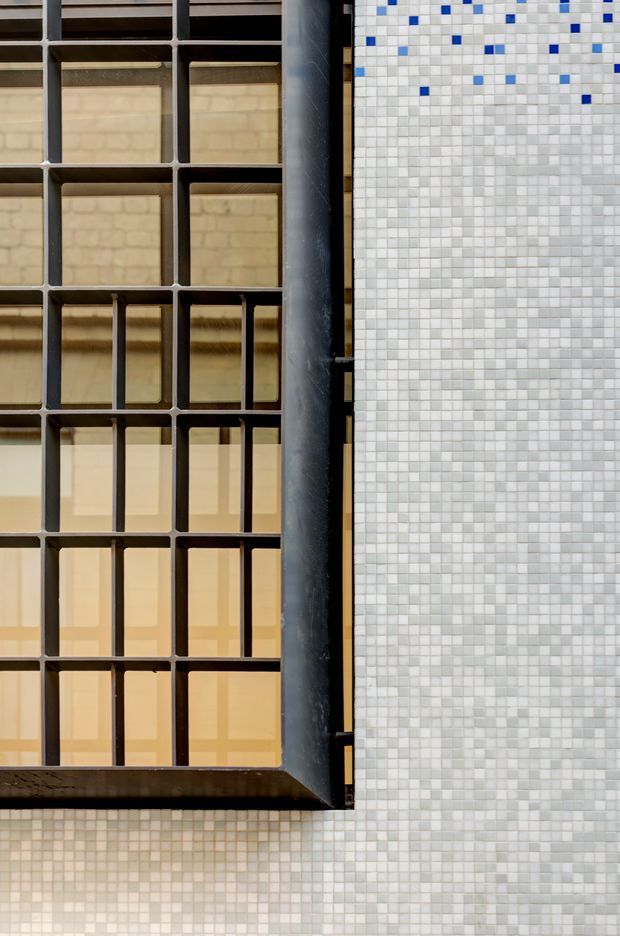 Close-up architectural detail showing a dark metal gridded window or screen beside a wall clad in small square mosaic tiles. The glazing reveals warm-toned interior surfaces behind the geometric frame, while scattered blue tiles create a subtle accent near the upper portion of the tiled wall. The composition is modern and highly graphic, with strong vertical and rectangular lines.