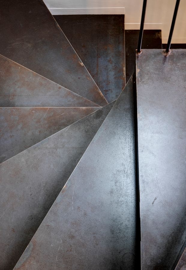 Close-up, top-down view of a sculptural spiral stairway with wedge-shaped dark metal treads radiating around a central point. A slim black metal handrail and balustrade are visible on the right, with pale walls in the background. The image emphasizes industrial materiality, geometric form, and the stair construction.