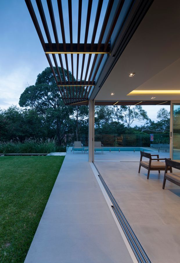 A contemporary indoor-outdoor living area opening onto a swimming pool and landscaped garden. Large retractable glazed doors create a seamless transition between the pale tiled interior and exterior terrace, while a projecting timber pergola provides strong linear shade overhead. The space is photographed at dusk, with recessed ceiling lights and warm concealed lighting contrasting against the blue evening light and surrounding trees.