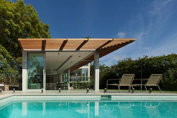 Contemporary poolside pavilion with a flat timber slatted pergola roof, slender structural columns, extensive floor-to-ceiling glazing, and a clean open-plan interior. A turquoise swimming pool dominates the foreground, with lounge chairs beside a landscaped hedge and mature trees in the background.