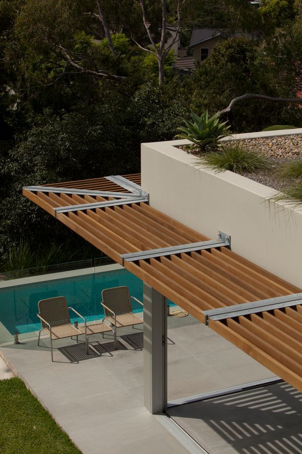A landscaped residential pool area viewed from above, featuring a cantilevered timber pergola or louvred shade structure with exposed metal supports. Below it is a turquoise swimming pool, pale concrete paving, and two outdoor lounge chairs. A raised cream rendered planter wall with pebble landscaping and succulents borders the terrace, with dense established vegetation surrounding the property.