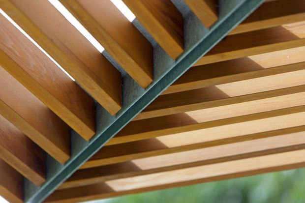 Close-up upward view of a timber-framed roof or pergola structure, with repeated warm-toned wooden rafters and slats supported by a dark grey-green exposed steel beam. Greenery is softly visible beyond the structure.