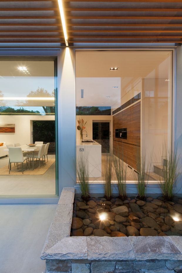 A contemporary indoor-outdoor architectural view showing a dining area and kitchen through full-height glazing. The kitchen features extensive dark timber wall cabinetry and a pale island, while the dining room has a light table and upholstered chairs. A covered exterior ceiling with exposed timber battens and integrated linear lighting frames the view. In the foreground, a raised stone-edged pebble garden with grasses and concealed uplights creates a landscaped courtyard feature.