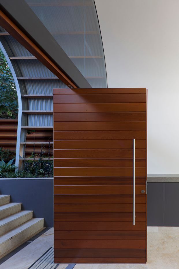 A close-up architectural view of a tall horizontal timber-panel pivot door with a long slim metal pull handle. The door is set within a contemporary entry area featuring pale stone steps and flooring, dark built-in cabinetry, exposed steel framing, corrugated translucent roofing or glazing, and planted landscaping visible beyond. The composition highlights the entry door, material detailing, and structural elements.