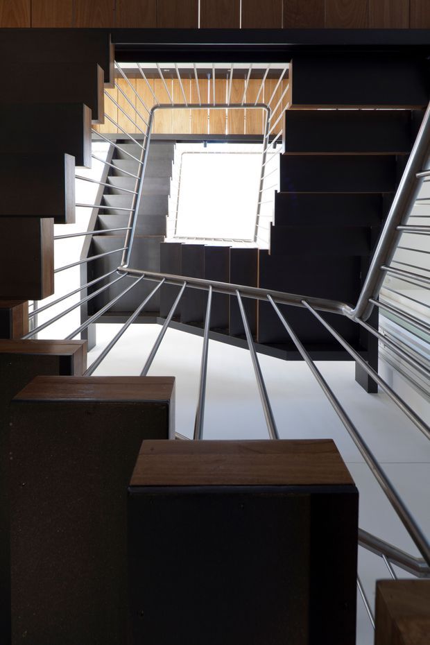 Upward-looking view through a compact modern stairway or landing, framed by dark timber-clad walls and built-in box-like joinery. Slim silver metal balustrades and handrails create strong linear geometry around a bright central skylight or roof opening, with additional timber ceiling lining visible above.