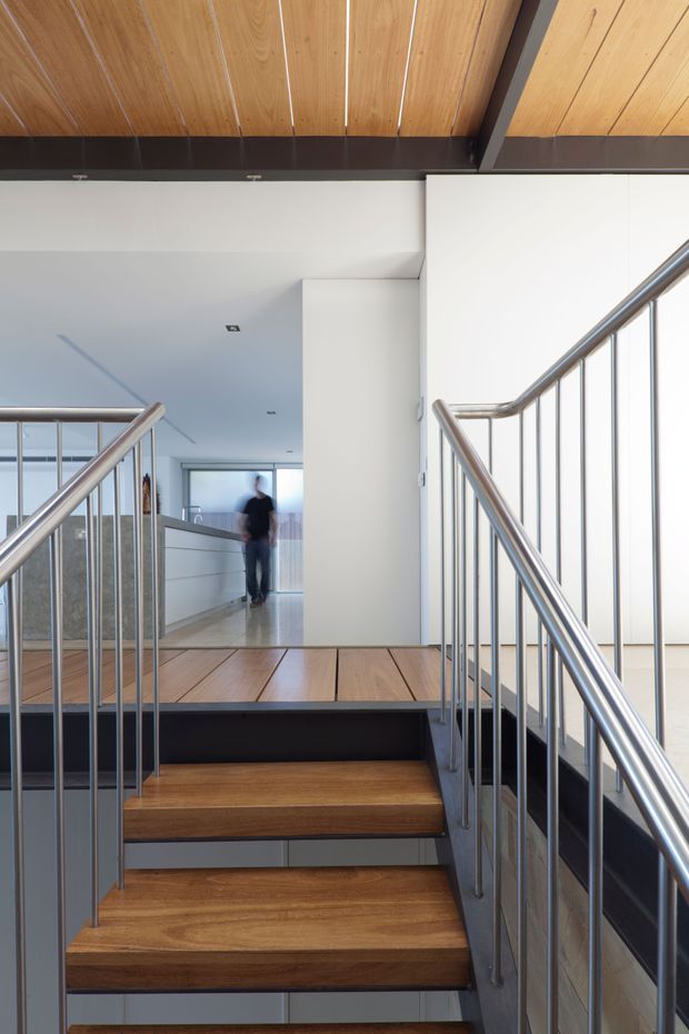 A bright modern stairway and landing viewed from the lower steps. The staircase has warm timber treads and matching timber-clad ceiling panels, dark structural framing, and slim stainless-steel balustrades with rounded handrails. White walls and a minimalist corridor extend beyond the landing, with a blurred person in the background.