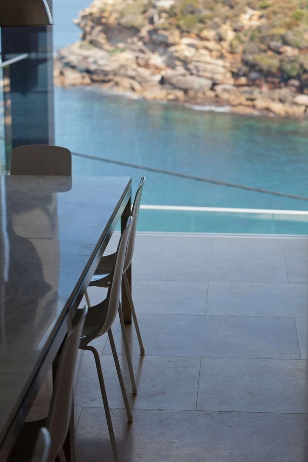 A contemporary dining area beside expansive glazing, with a long dark dining table and streamlined chairs on large pale stone tiles. The glazing opens to a dramatic waterfront outlook with clear blue water and rocky shoreline, creating a minimal, refined indoor-outdoor setting.