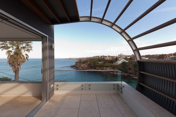 A covered coastal terrace or balcony overlooking a sheltered blue-water bay and rocky headland. The space features large pale stone or porcelain floor tiles, a frameless glass balustrade, dark metal cladding and framing, and a distinctive curved translucent roof canopy with exposed radial beams. Sliding glazing opens toward the view, with a palm tree and neighbouring waterfront buildings visible beyond.