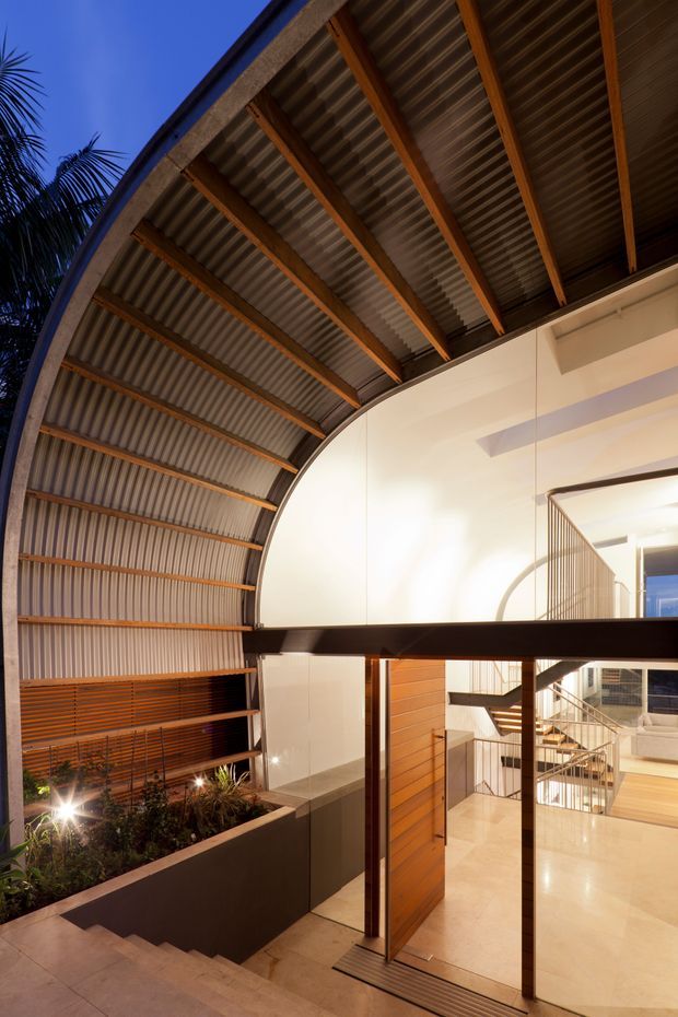 A dramatic modern interior atrium beneath a large curved corrugated-metal roof, with exposed timber rafters, a mezzanine or upper-level walkway, open staircases, glass balustrades, warm timber wall cladding, polished pale flooring, and an indoor planted courtyard. The space is illuminated warmly against a deep blue evening exterior visible through the opening.