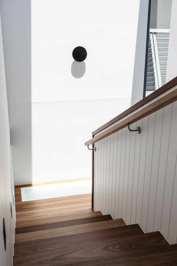 A bright contemporary stairway viewed from above, with dark timber treads, a white vertical-panel balustrade, and a simple timber handrail supported by dark metal brackets. The stair descends toward a landing with a rectangular skylight or glazed opening, while a black circular wall light punctuates the smooth white wall. A louvred window is visible at the upper right.