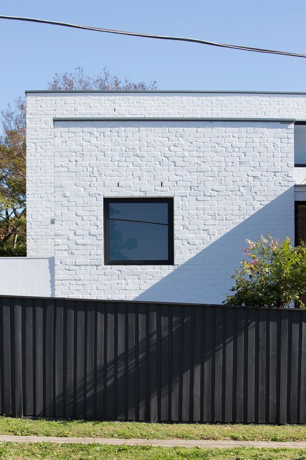 Exterior view of a compact contemporary building with a flat roof, white-painted textured brick walls, dark-framed windows, and a tall black vertical-panel boundary fence. Strong sunlight creates a diagonal shadow across the facade, with trees and shrubs partially visible around the property.