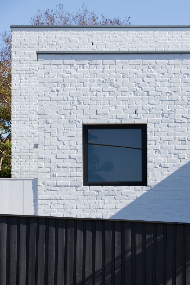 Close-up exterior view of a compact, flat-roofed building facade with white-painted exposed brickwork, a dark-framed rectangular window, and dark vertical-clad fencing or screening in the foreground. Strong sunlight creates sharp shadows across the wall; leafless tree branches are visible behind the building.