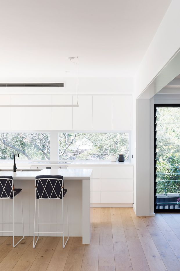 Bright, minimalist white kitchen with a large island and two black-and-white bar stools. The kitchen features full-height white cabinetry, a pale stone or engineered-stone benchtop, light timber flooring, a slim linear pendant, and expansive windows overlooking leafy trees. A black-framed louvred glass door opens to an exterior balcony or deck.