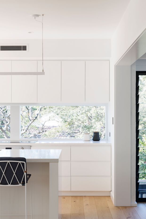 Bright minimalist kitchen with extensive white flat-panel cabinetry, a pale stone island benchtop, integrated appliances, and a large horizontal window overlooking leafy trees. A black-and-white bar stool sits at the island, while a dark-framed glazed door or window is visible at right. Light timber flooring and a simple linear pendant reinforce the clean contemporary aesthetic.