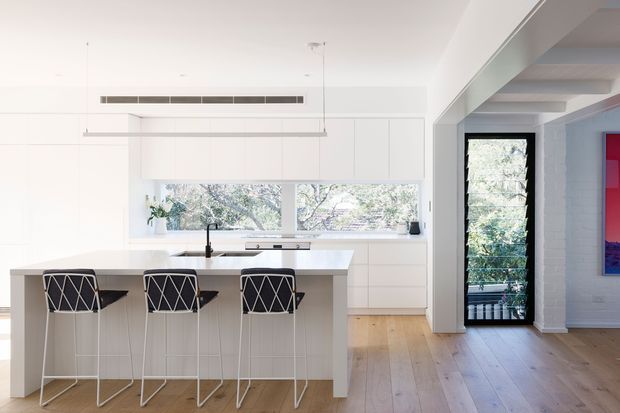 Bright open-plan kitchen with a large white island and three bar stools, extensive white flat-panel cabinetry, integrated appliances, and a black tap. Light timber flooring runs through the space. Large windows above the benchtop overlook leafy vegetation, while a wide opening leads to an adjacent room with exposed white-painted brick and ceiling beams. The interior is clean, minimalist, and highly illuminated by natural light.