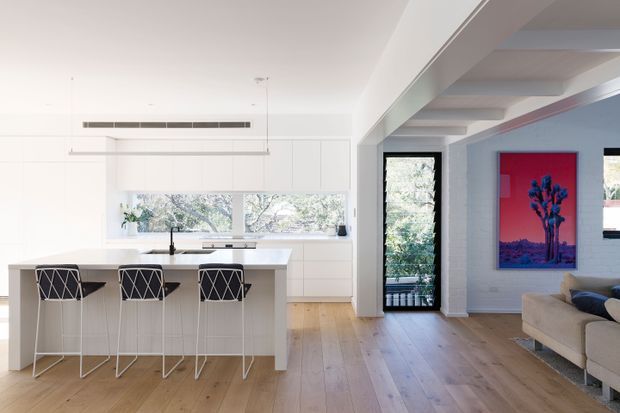 Bright open-plan interior showing a minimalist white kitchen with a large island and three bar stools, opening directly into a living area. The kitchen features flat-panel white cabinetry, a white benchtop, integrated appliances, a slim linear pendant light, and extensive windows overlooking trees. The adjoining living space has pale timber flooring, exposed white ceiling beams, a narrow glazed door, and a vivid red and blue artwork.