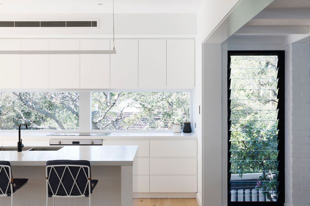 Bright contemporary kitchen with white full-height cabinetry, a white island with integrated sink and black tapware, and two black-and-white stools. Large horizontal windows overlook leafy greenery, while a black-framed louvred window and exposed white brick wall are visible through the wide opening on the right. Pale timber flooring and simple linear pendant lighting create a clean, airy interior.