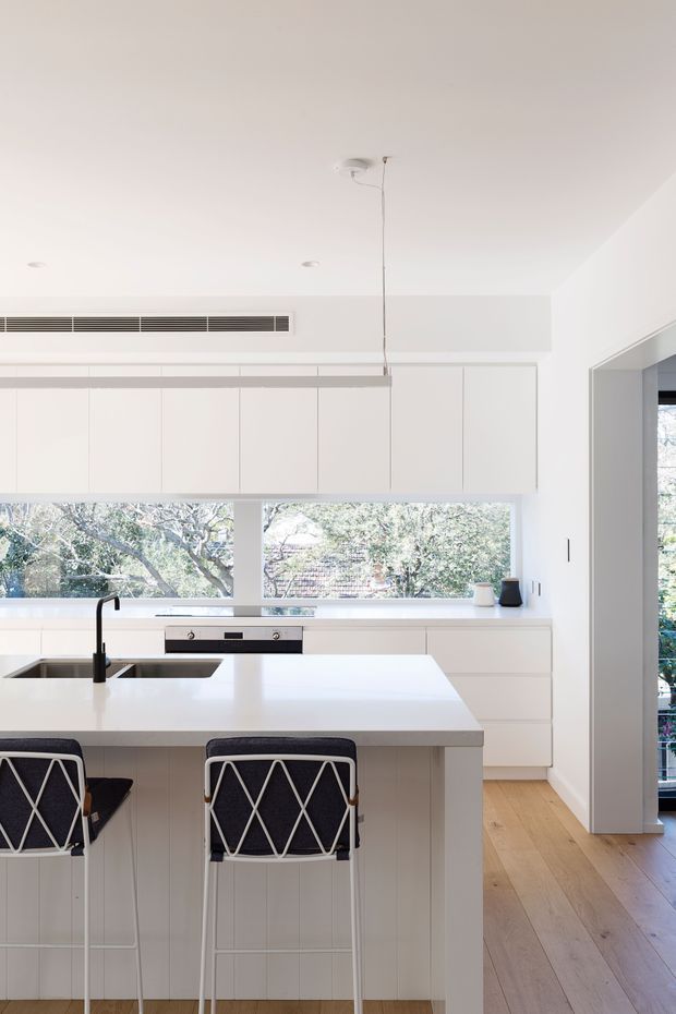 Bright, minimalist contemporary kitchen with extensive white flat-panel cabinetry, a large white island with an integrated sink and black tap, and two black-and-white bar stools. Pale timber flooring, recessed ceiling lights, a linear pendant, and broad windows overlooking leafy trees create an airy, clean interior.