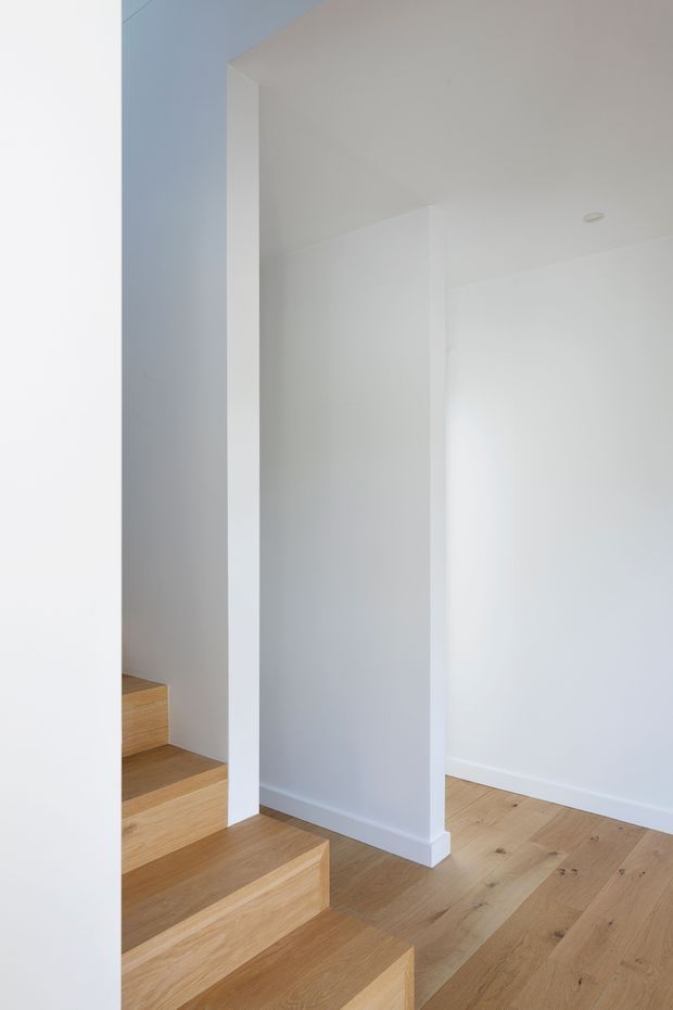 Minimalist interior view of a bright white stairwell and adjoining landing, featuring broad timber stairs and matching pale timber flooring. Clean white walls, a simple square structural column, white skirting, and a recessed ceiling light create a sparse contemporary composition.