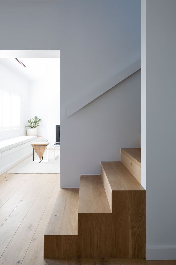 Minimalist interior view focused on a sculptural timber staircase with built-in stepped joinery and a simple white handrail. The stairs occupy the foreground beside a bright, white living area visible beyond, with pale timber flooring, a small side table, and a potted plant. The space has clean lines, abundant natural light, and restrained detailing.