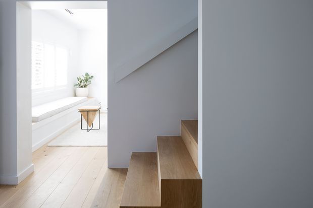 Bright minimalist interior showing a timber staircase with integrated stepped forms beside an airy window seat. Pale timber flooring, white walls, simple white cabinetry or seating, and a small sculptural side table create a clean, restrained composition.