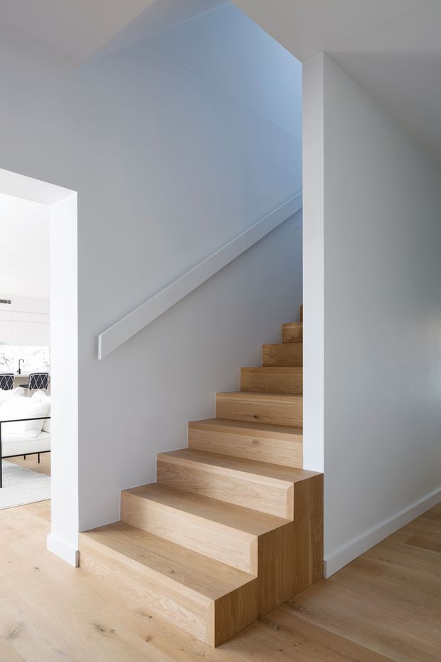 A minimalist interior stairway with broad solid timber steps and matching timber flooring, set against smooth white walls and a simple white handrail. The staircase rises beside an open doorway into a bright living area visible at left, with clean-lined furniture and abundant natural light. The composition emphasises the sculptural stair form, pale wood, and restrained detailing.