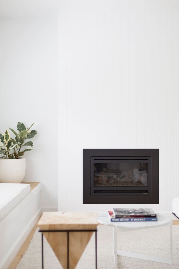 Bright minimalist living room with a wall-mounted black glass-front fireplace set into white vertical-panelled walls. A pale upholstered sofa, light timber coffee table, white side table with books, and potted plant create a clean, contemporary interior.